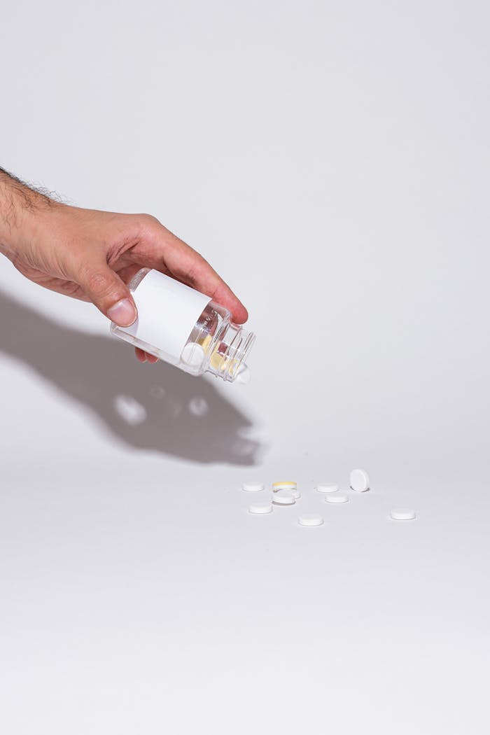 about-02 A hand holding a glass bottle pouring pills onto a white surface in a minimalist setup.
